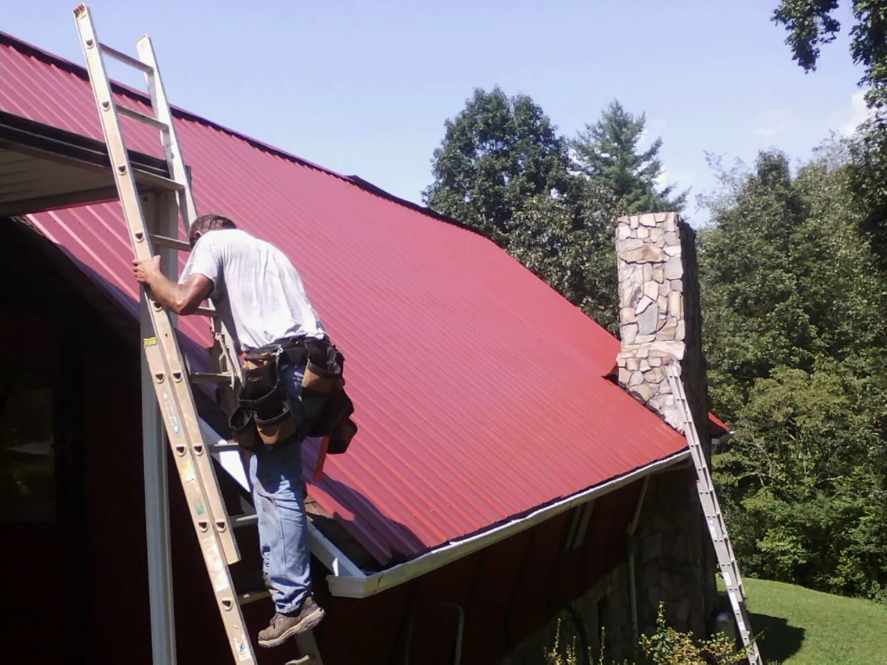 Roofer on a ladder beside a red metal roof and stone chimney — Soffit & Fascia Repair by Roofing Brothers in Mims